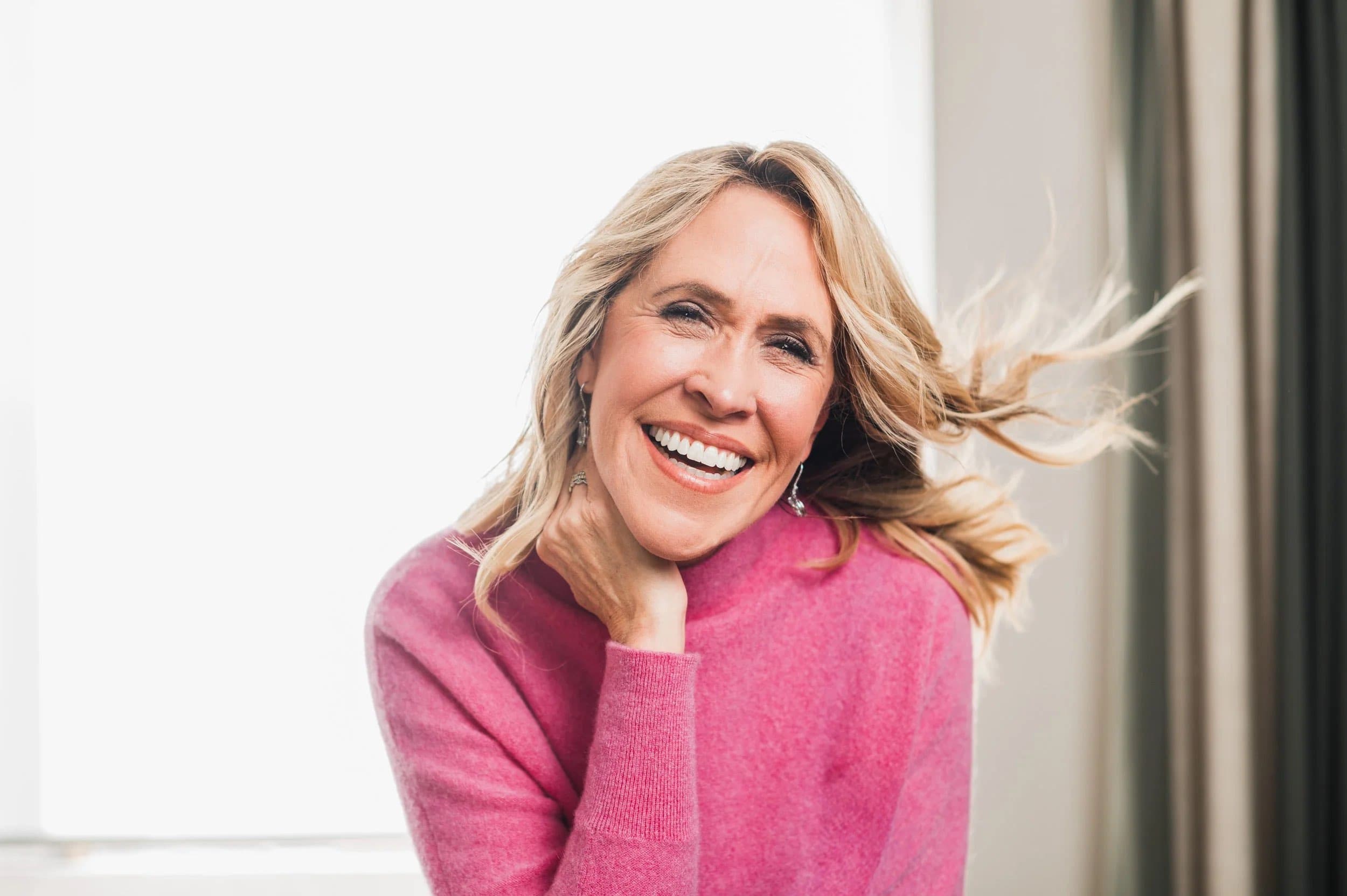 A laughing woman in a pink jumper against a white background, her hair in motion