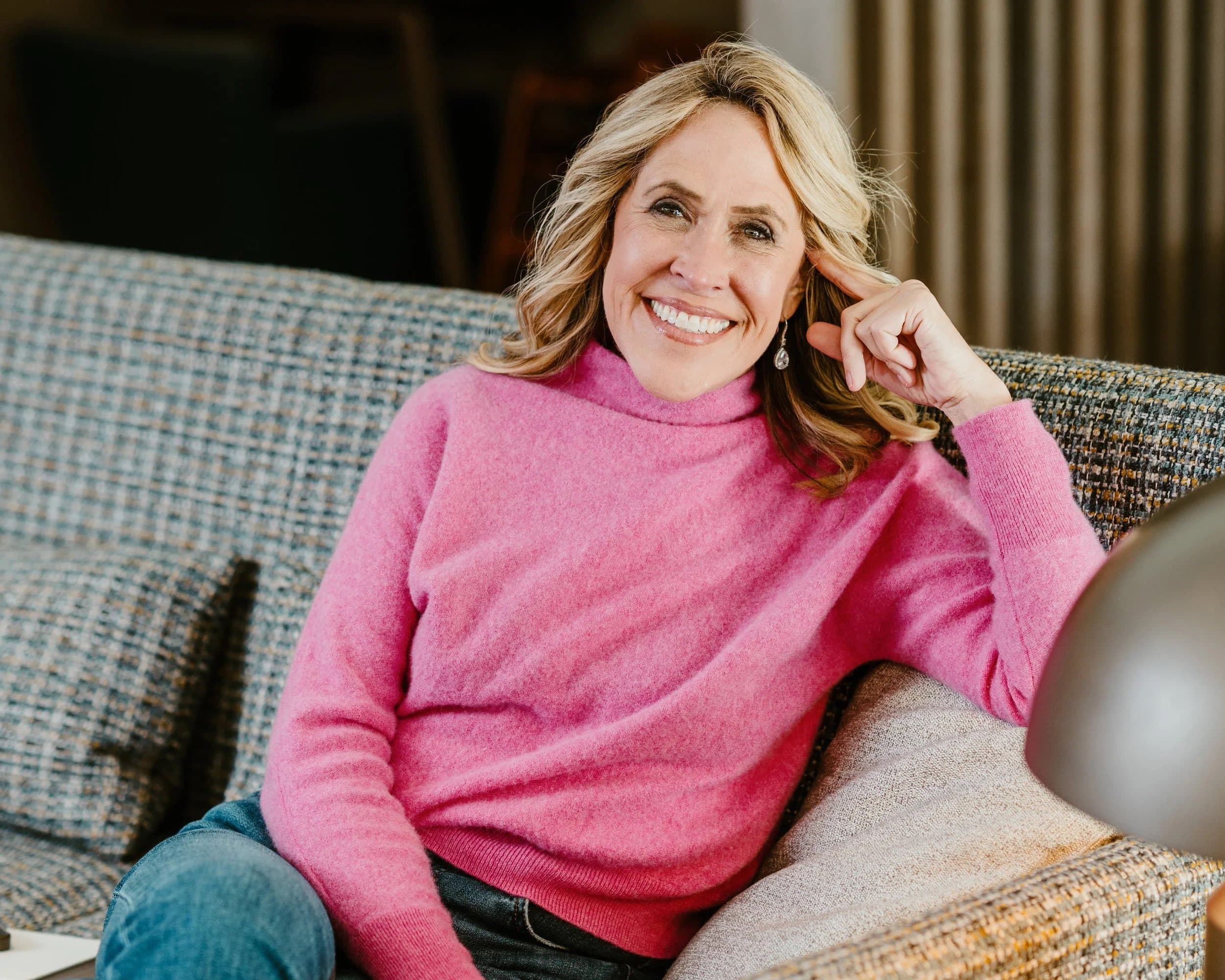 A smiling woman in a pink jumper resting her head on her hand on a sofa