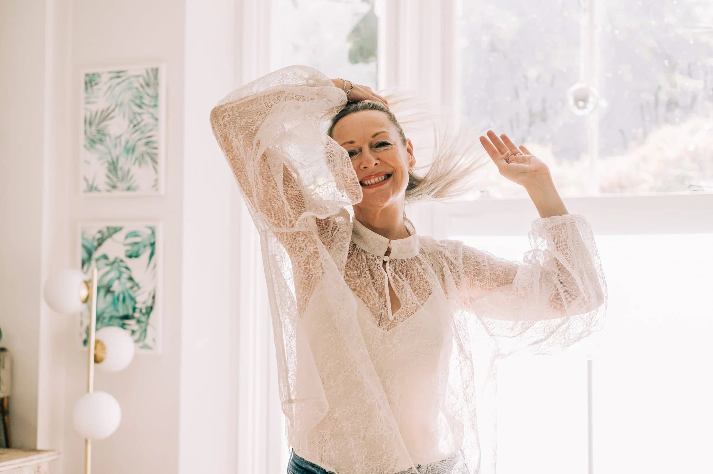 A woman in a white lace blouse laughing by a sunlit window, one hand raised