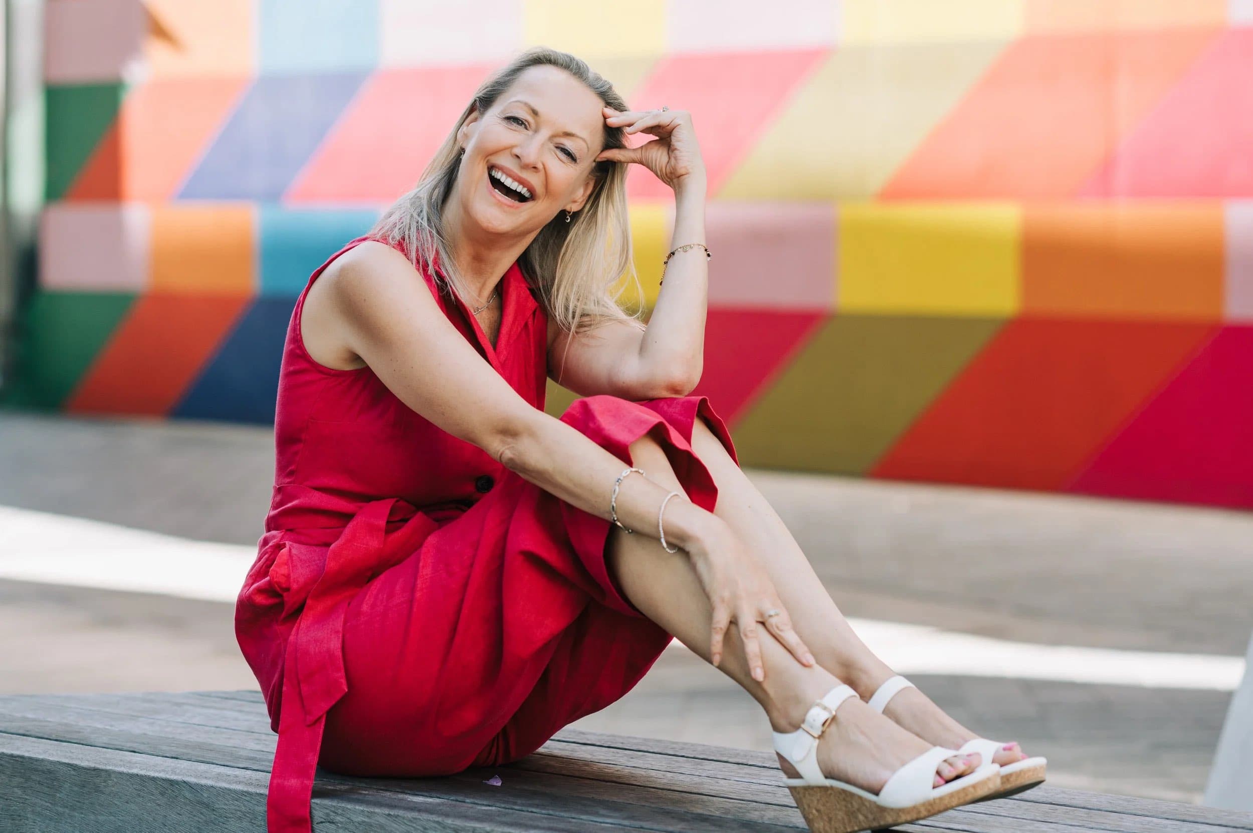 A laughing woman in a red dress sitting on decking in front of a brightly painted wall