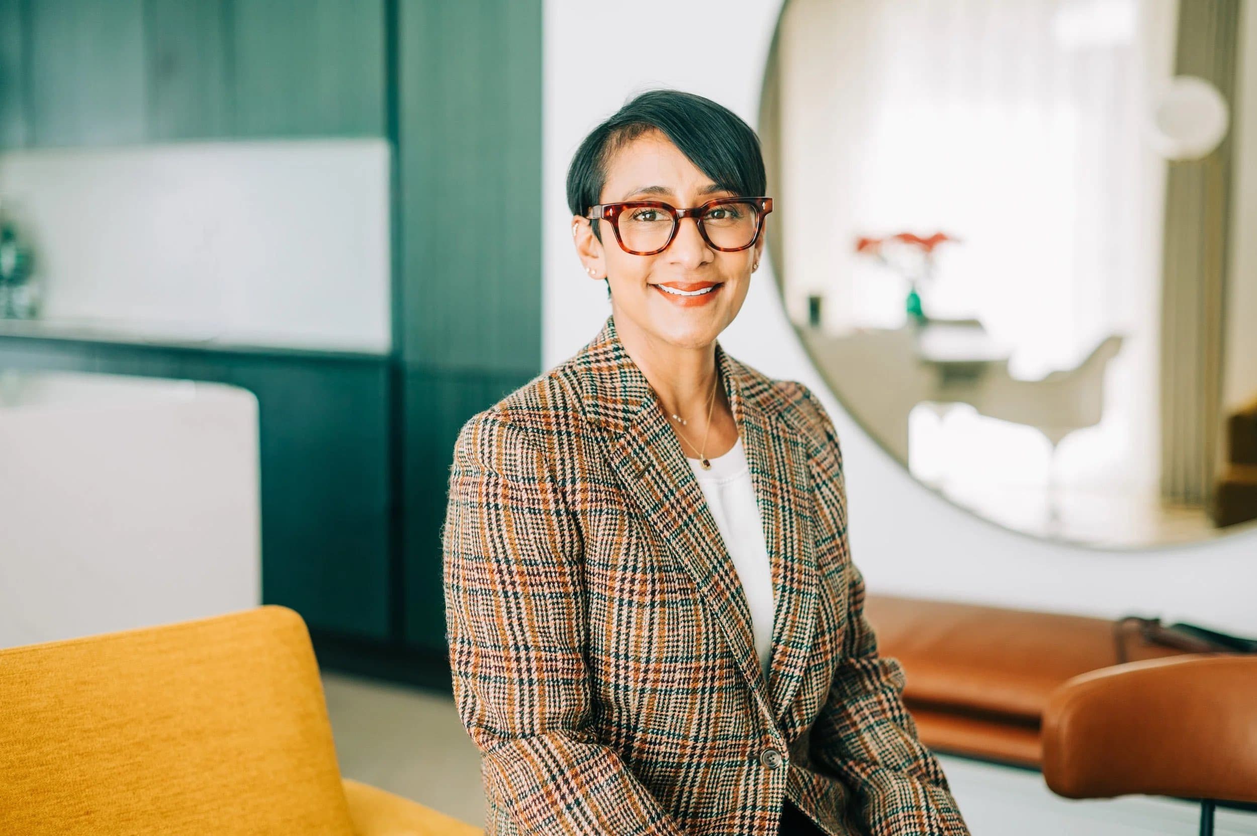A woman in glasses and a checked blazer seated in a bright office lounge