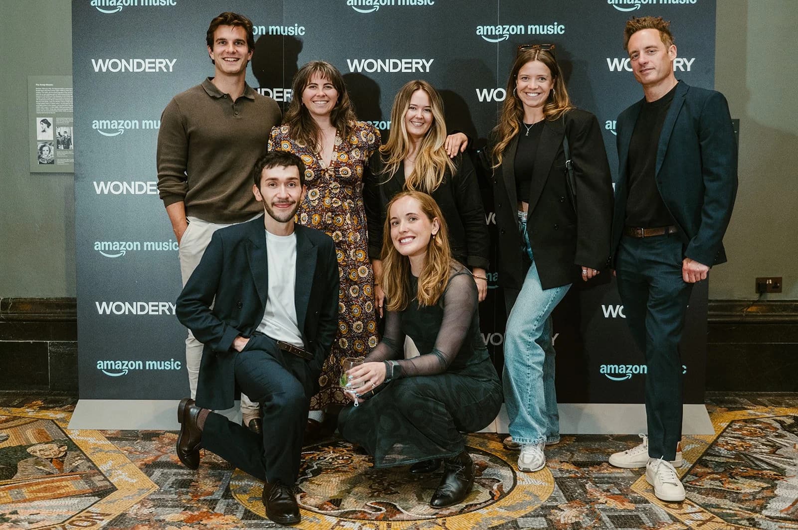 A group of seven posing in front of an Amazon Music and Wondery press wall