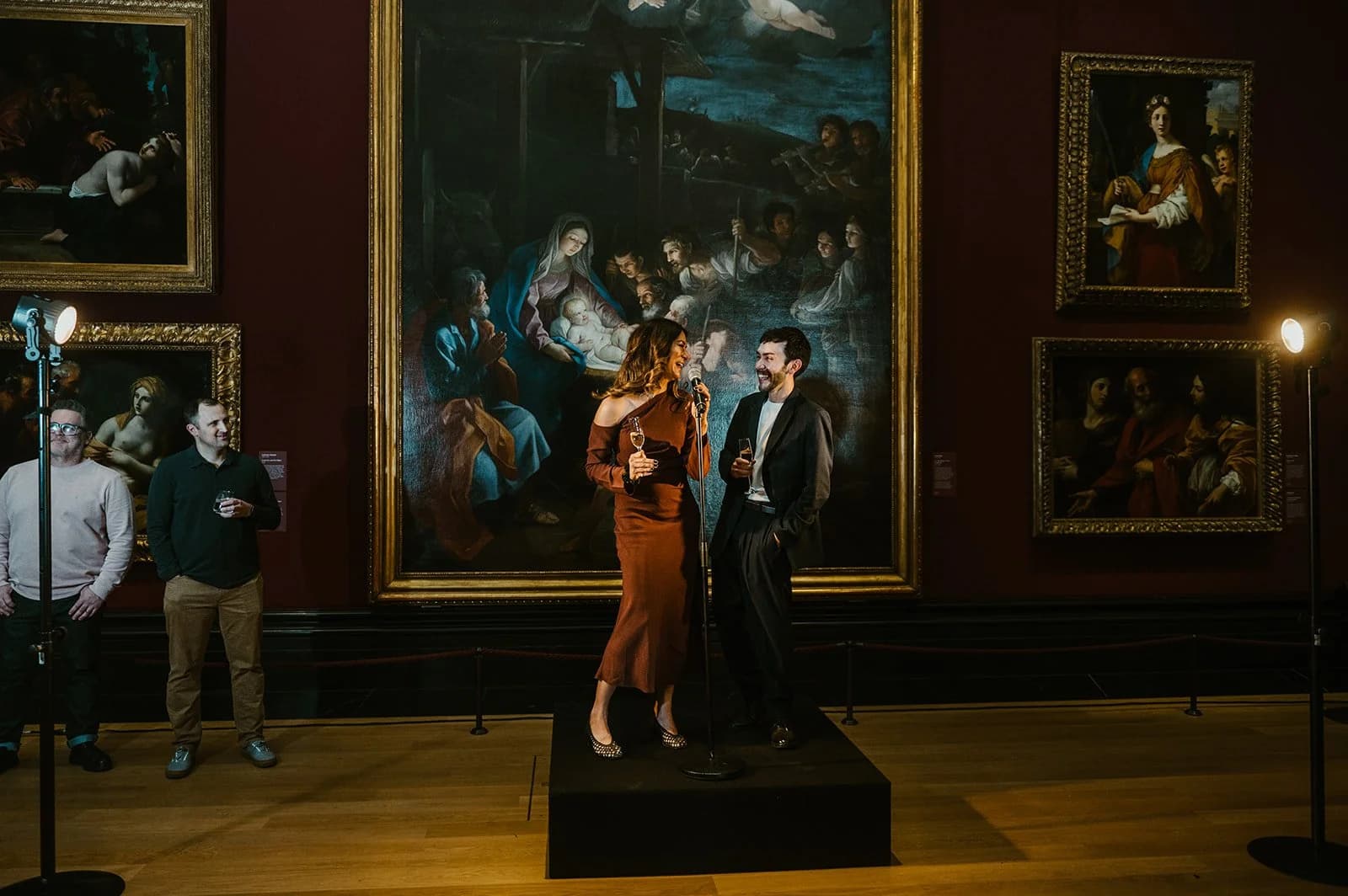 A woman and a man speaking on a small stage in a gallery of large oil paintings