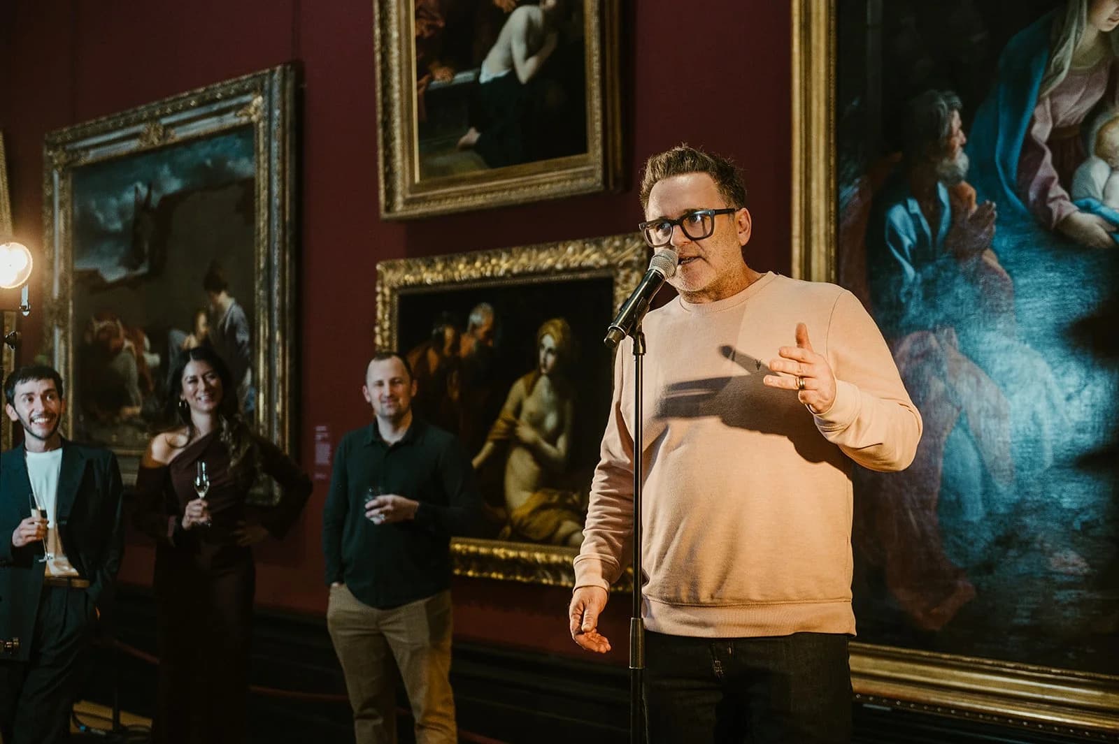 A man in glasses and a peach sweatshirt speaking into a microphone in a gallery of gilt-framed paintings