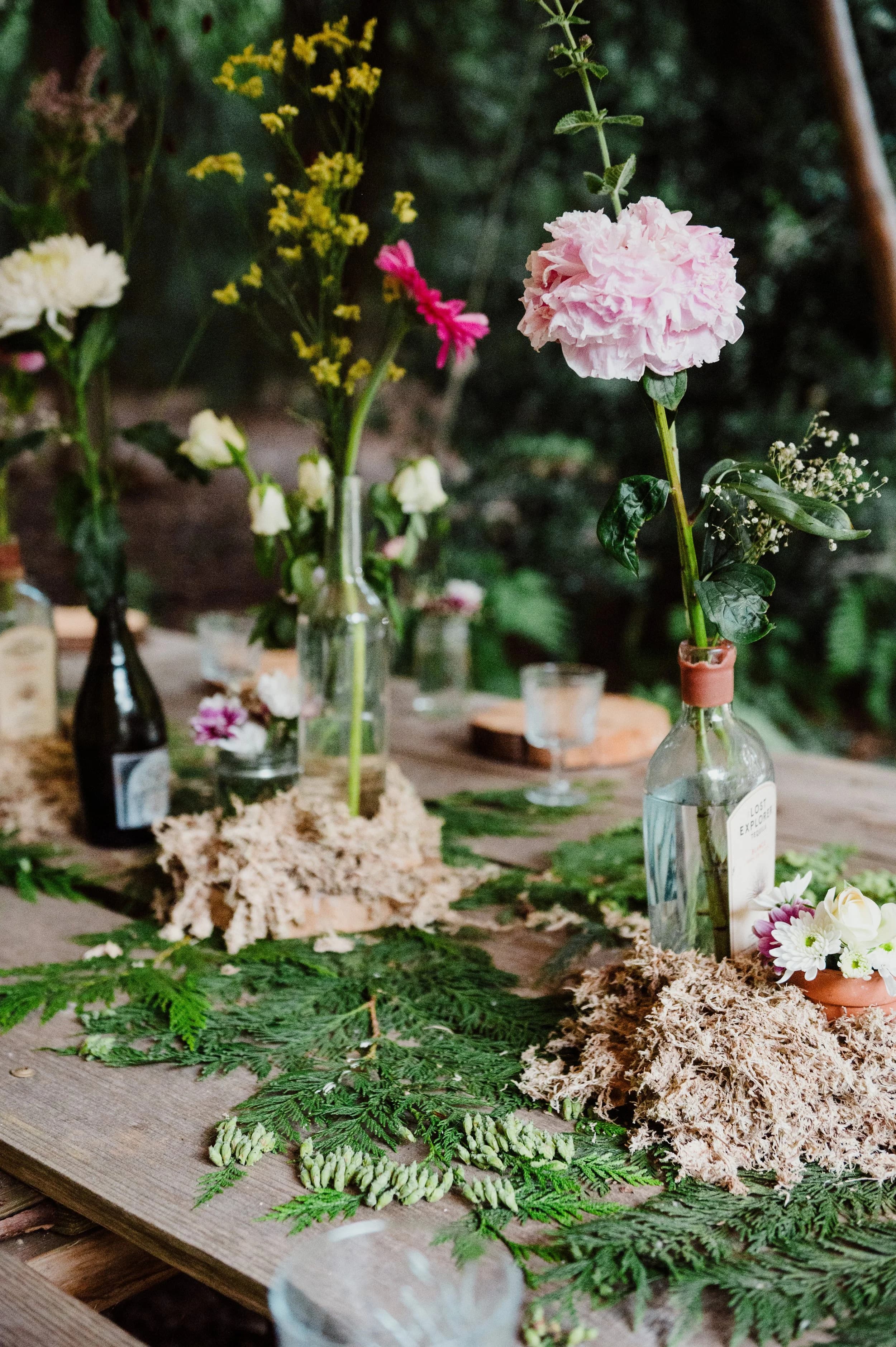 Table decorations of single stems in glass bottles among pine sprigs and moss on a rustic wooden table