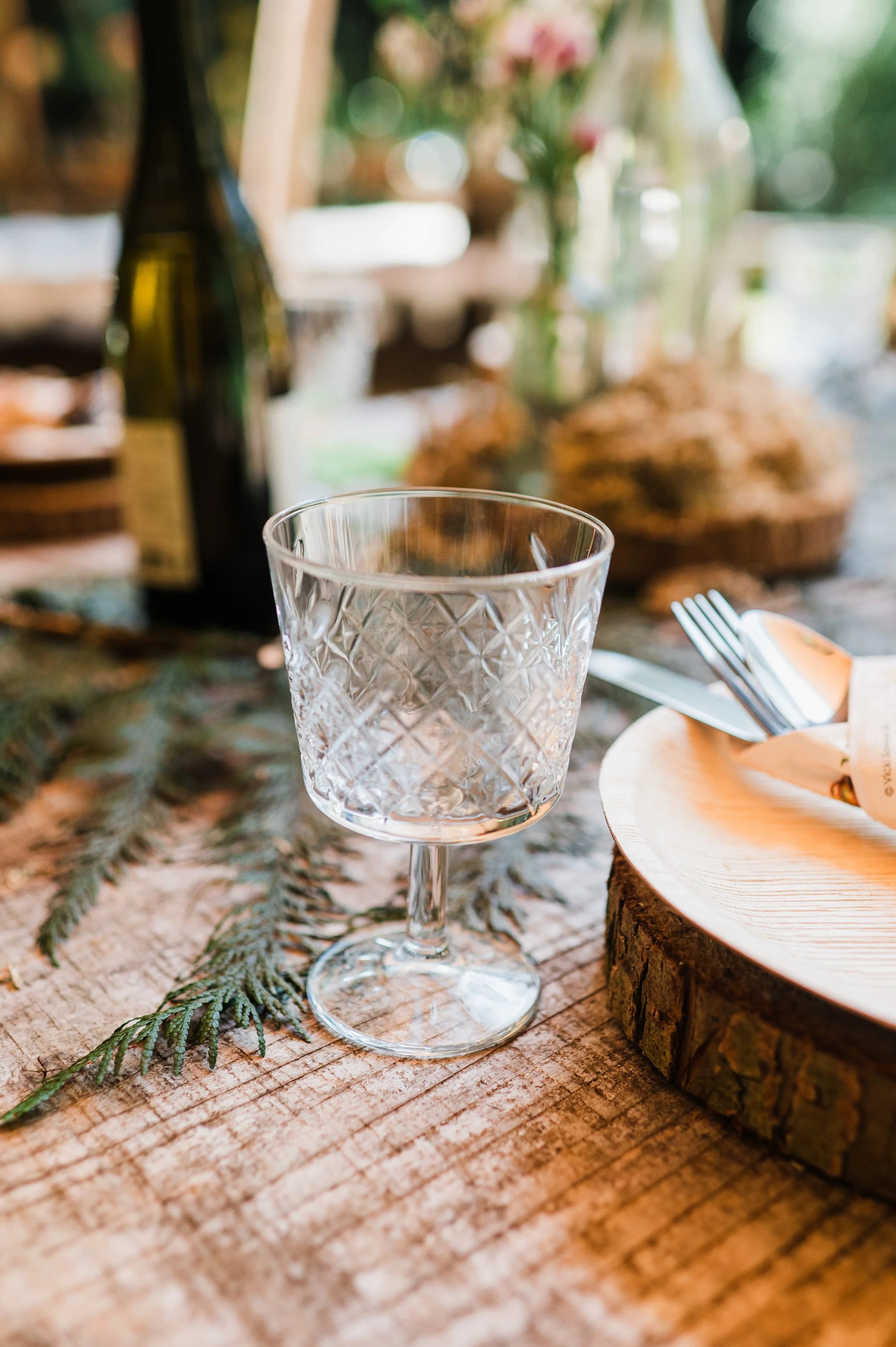A cut-glass goblet on a rough wooden table beside a wooden slice plate, cutlery and pine sprigs
