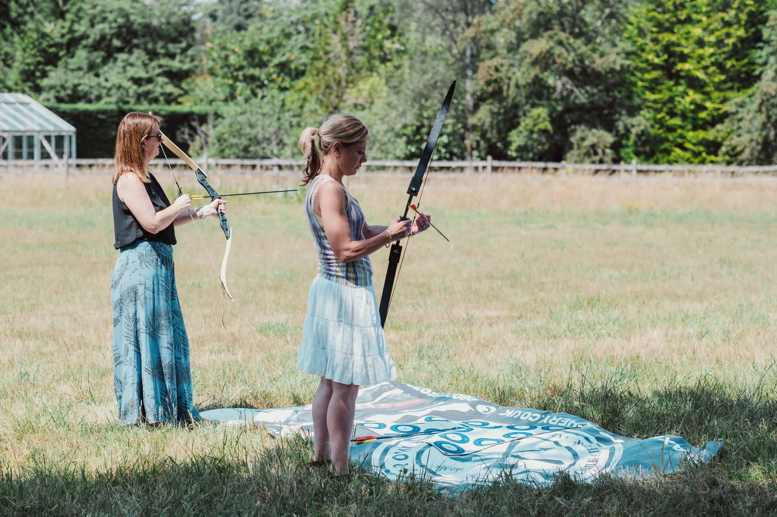 Two women with archery bows on the grass, one fitting an arrow while the other draws her bow