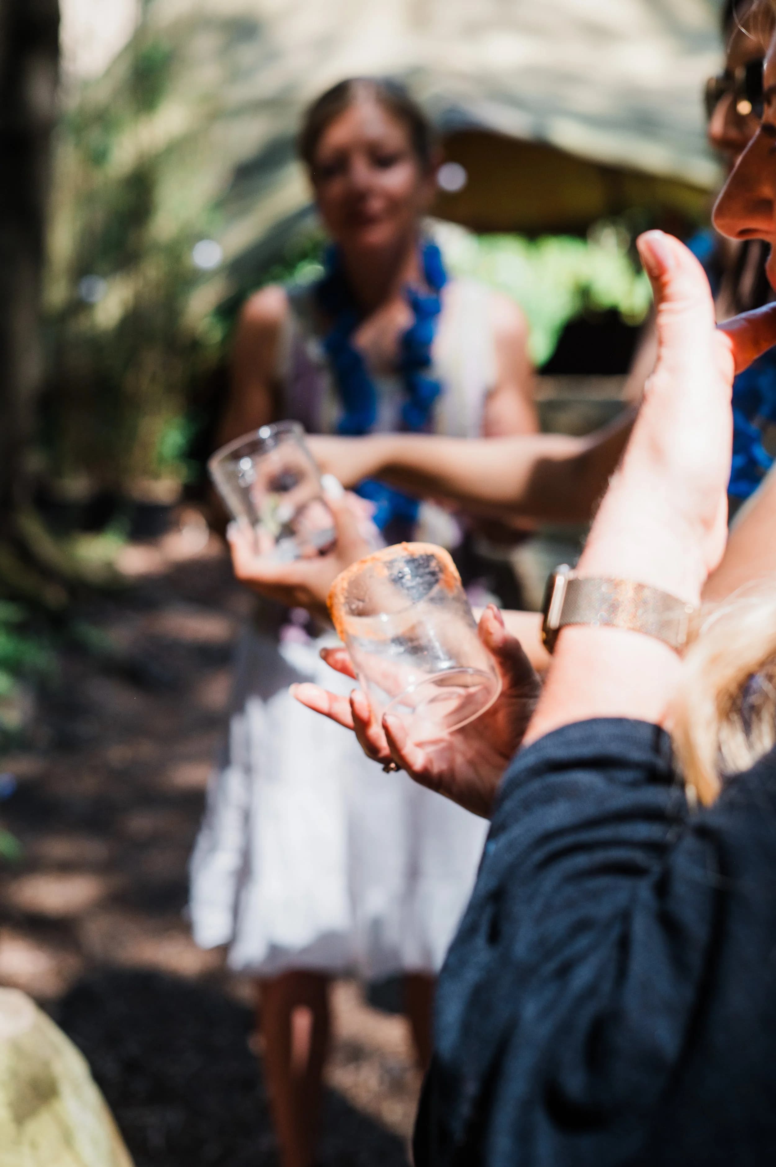 Guests in blue flower garlands holding small glasses together, with a woman smiling in soft focus behind