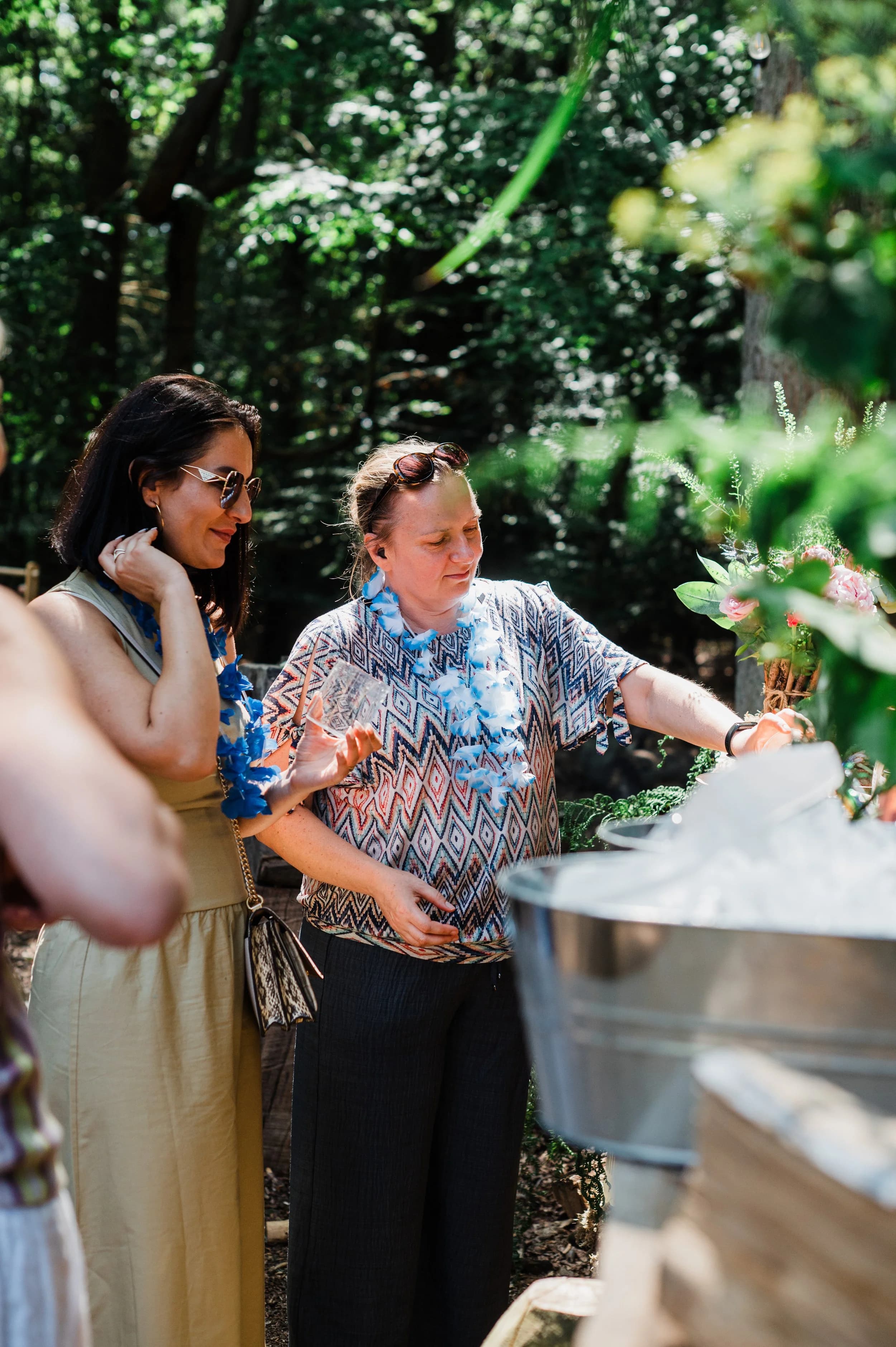 Two women in blue garlands chatting beside an ice bucket among trees at a woodland party