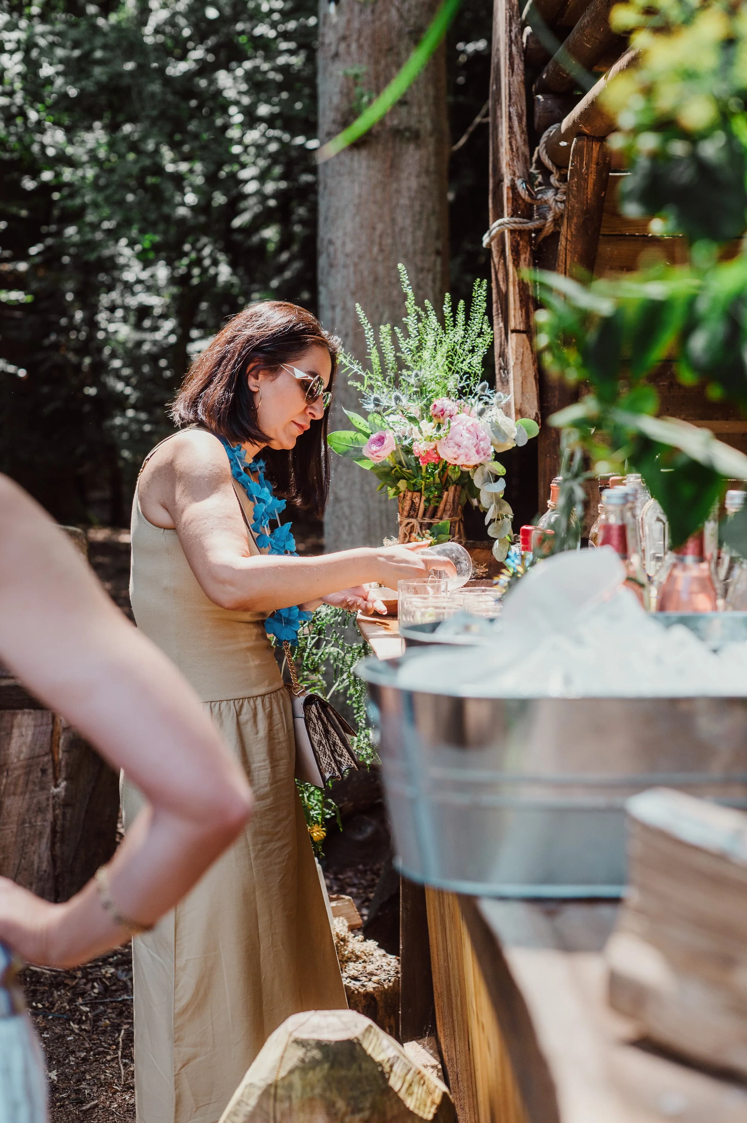 A woman in sunglasses and a blue garland preparing drinks at a rustic woodland bar