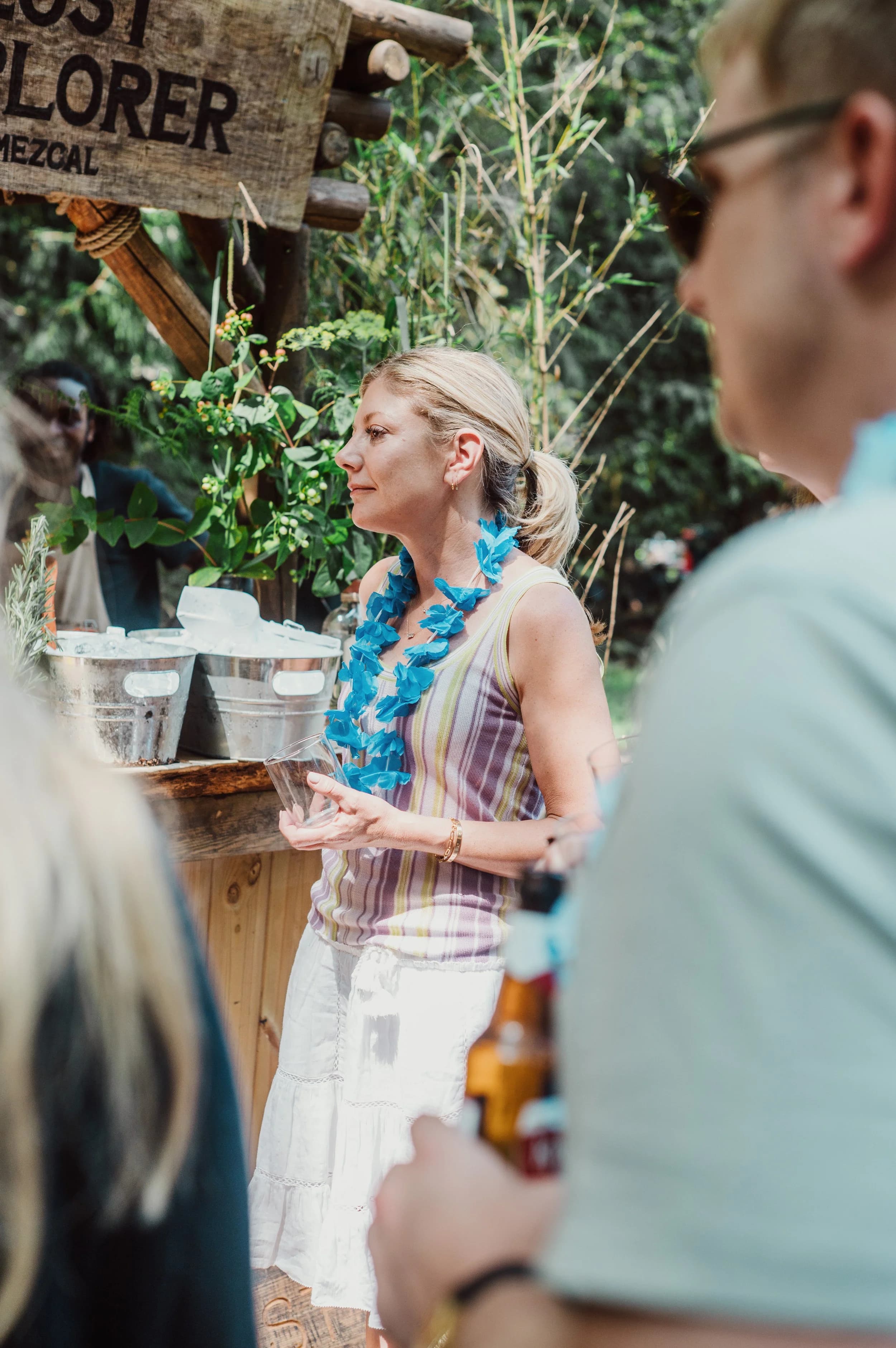 A woman in a blue garland holding a glass at a wooden bar, a beer in the foreground