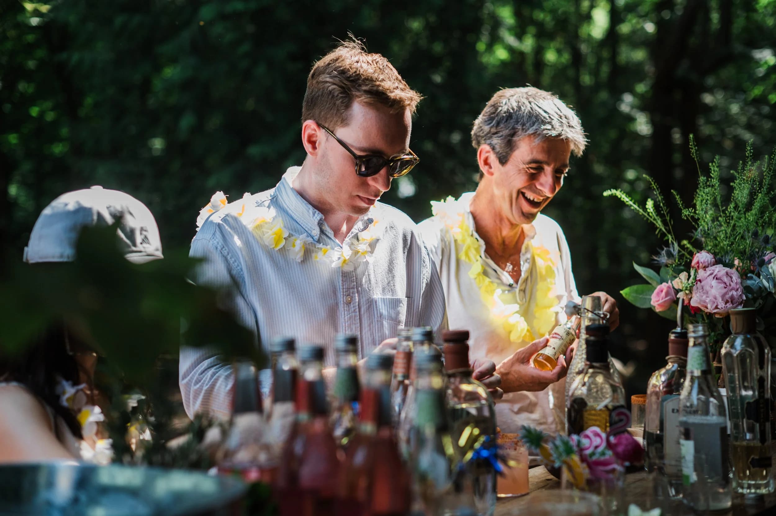 Two men in flower garlands laughing at an outdoor drinks bar among trees