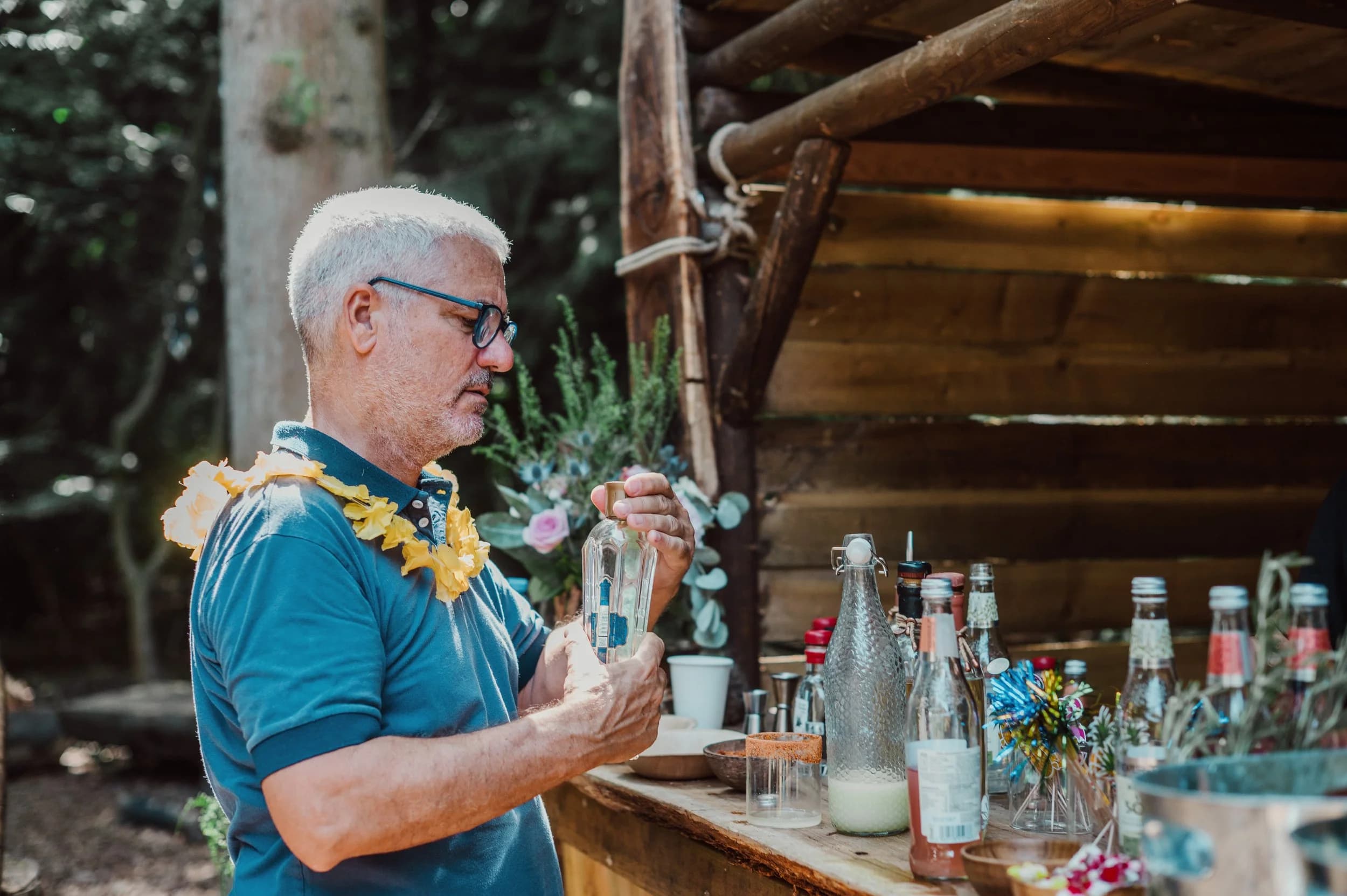 A grey-haired man in a teal polo and flower garland pouring a drink at a rustic wooden bar