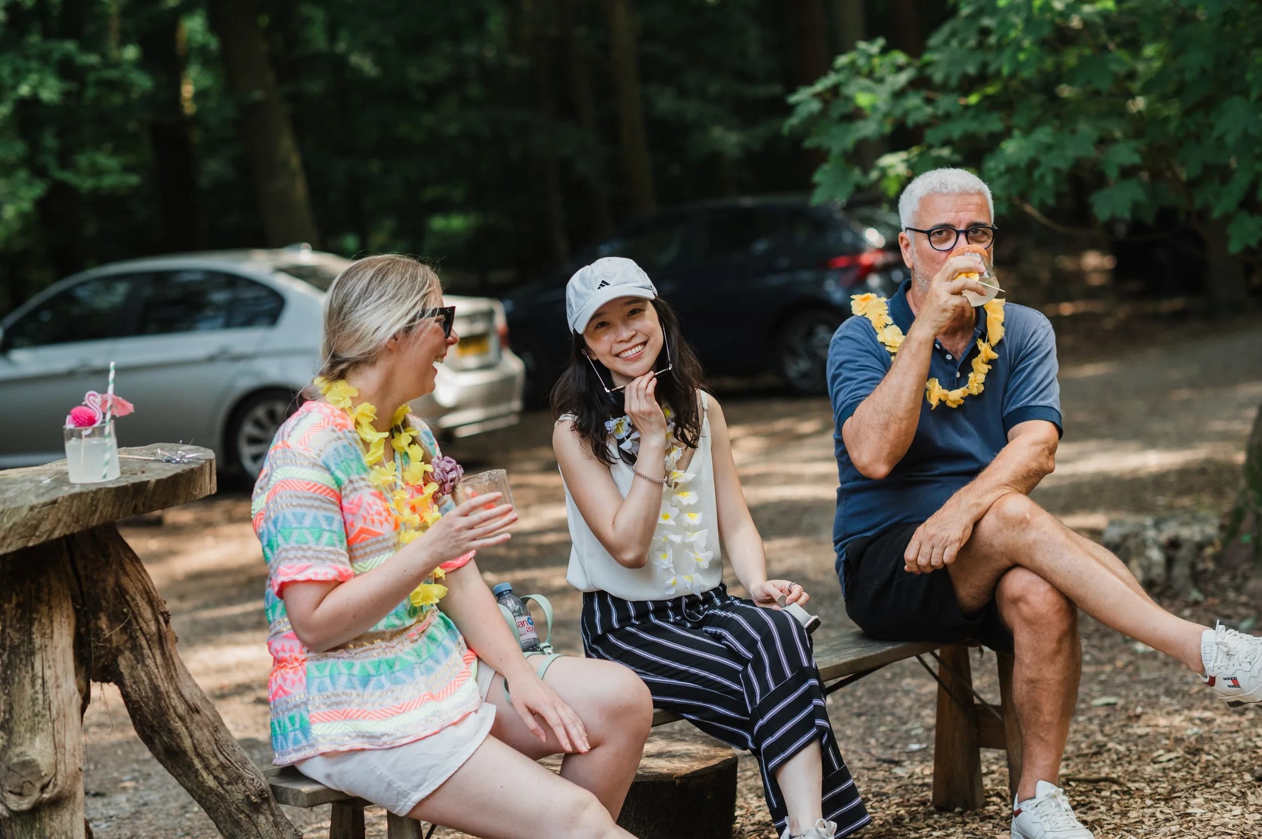 Three guests in flower garlands chatting on a log bench in a sunlit woodland clearing