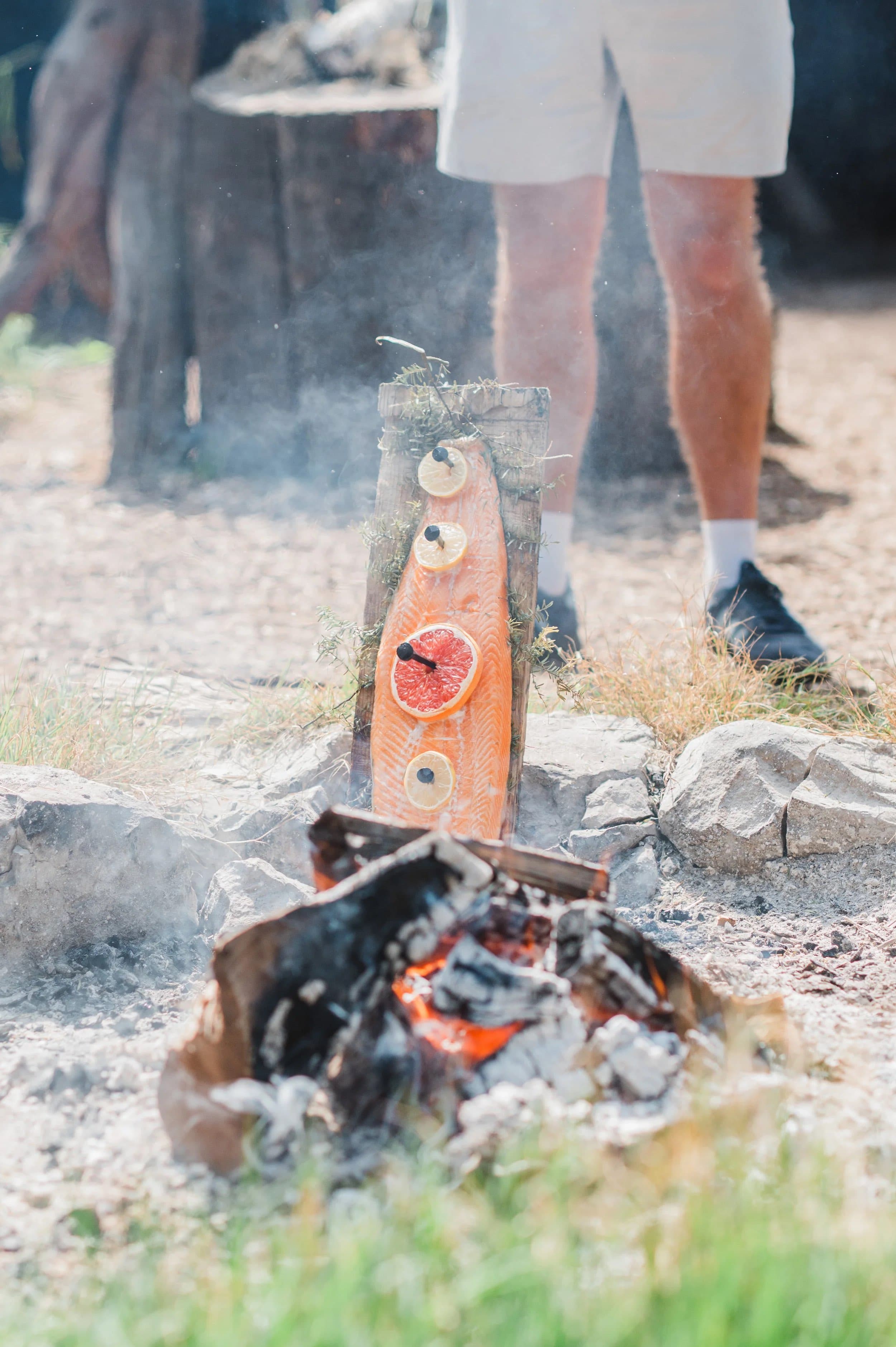 A salmon fillet on a wooden plank cooking beside an open fire, a guest standing behind