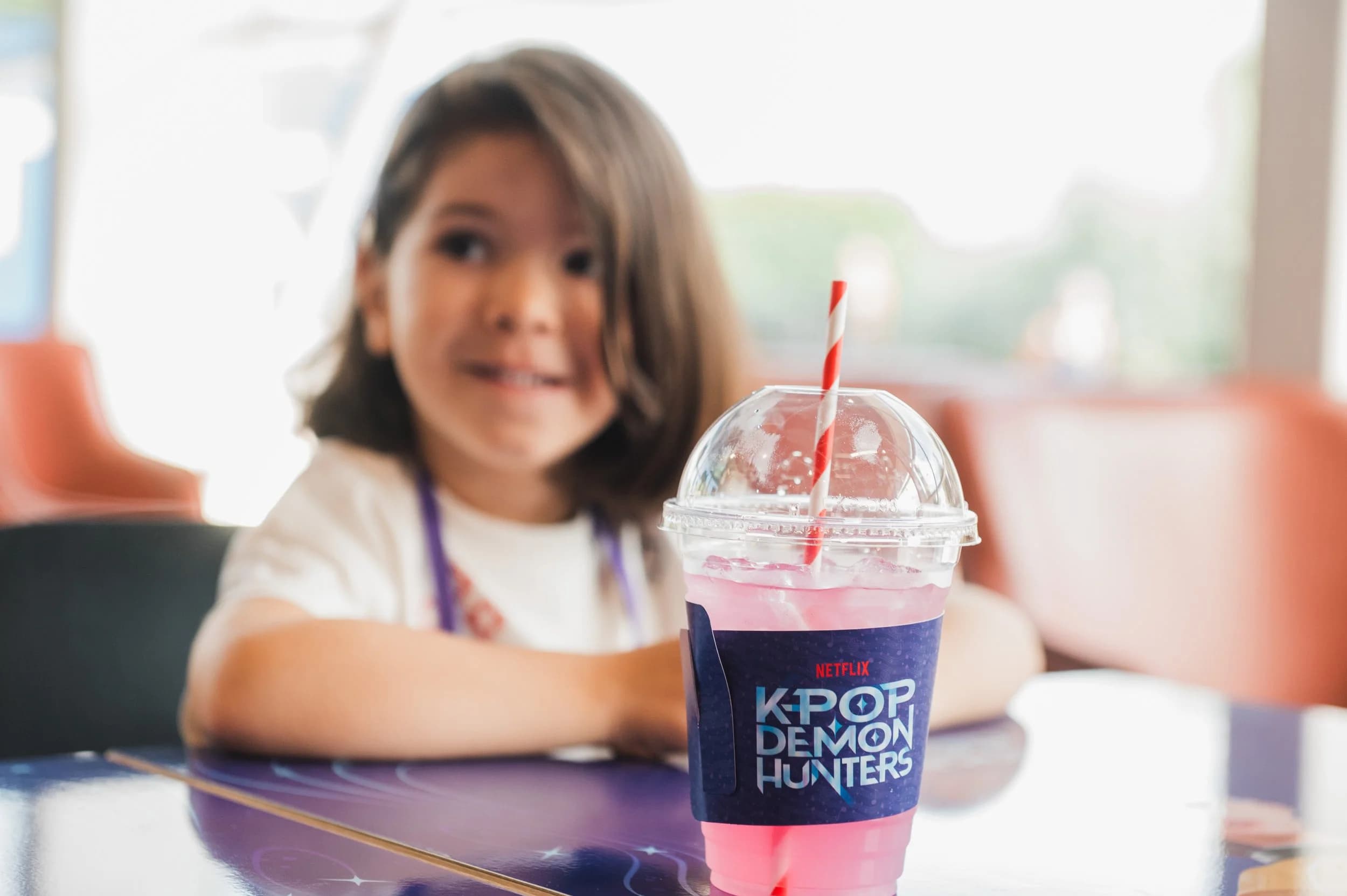 A smiling girl sitting behind a pink drink in a K-Pop Demon Hunters branded cup
