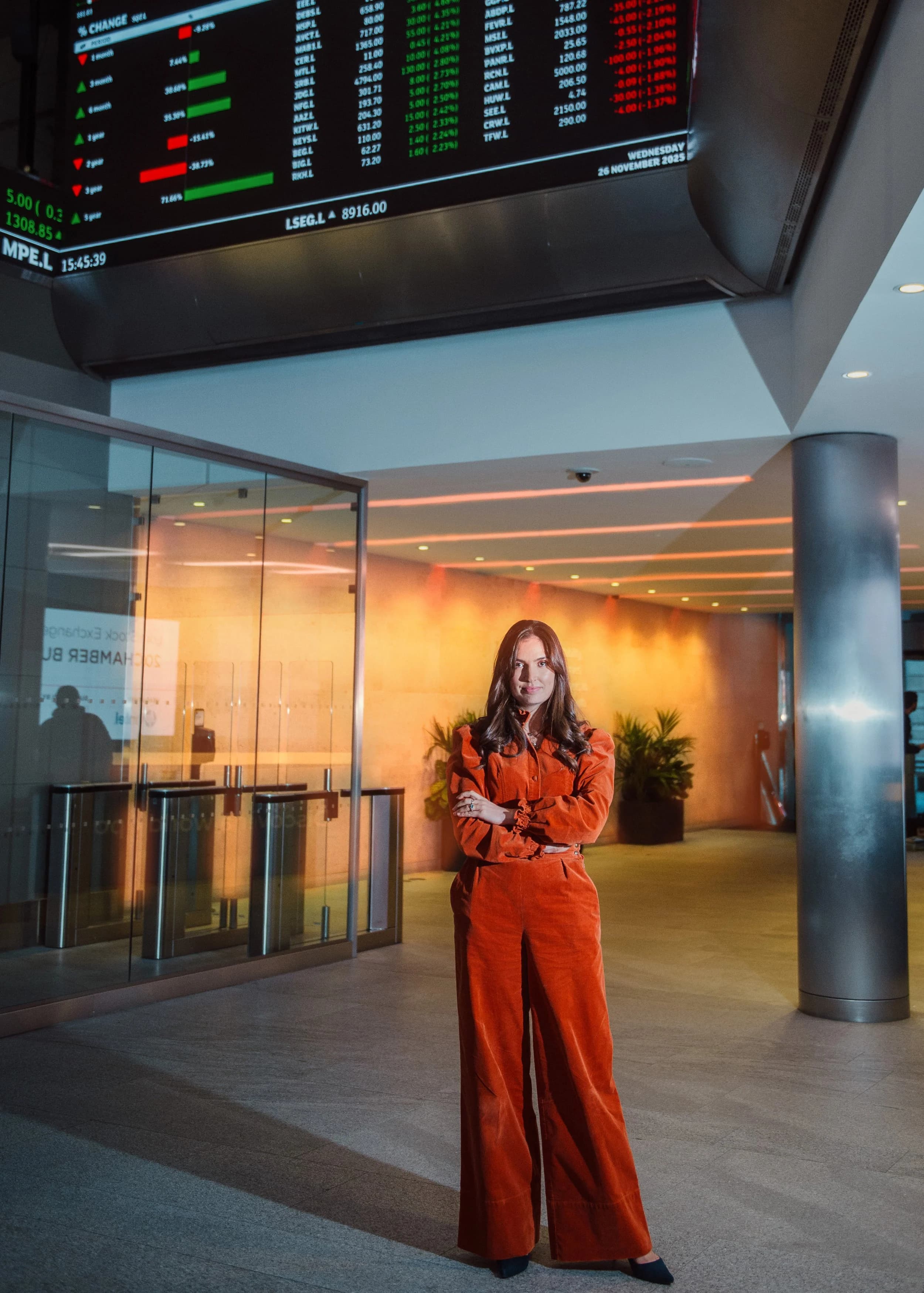 A woman in a rust-coloured suit standing in an office lobby beneath trading screens