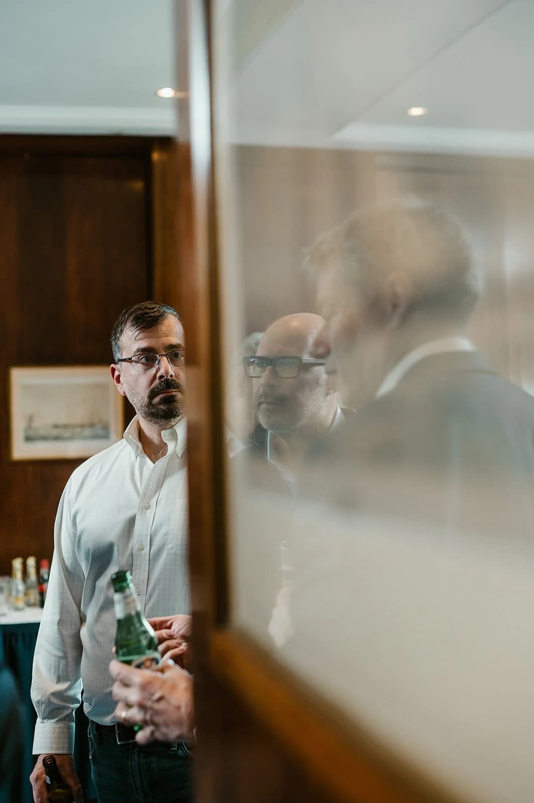 A man in a white shirt holding a drink, seen through a glass partition