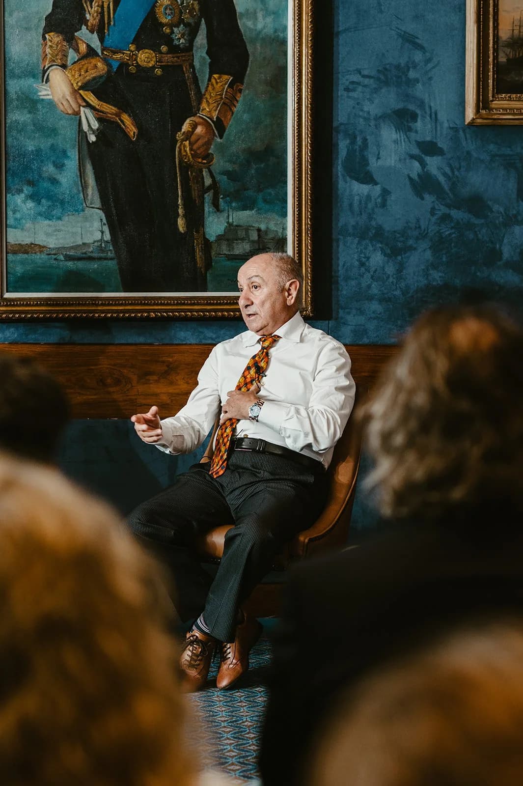 A man in an orange patterned tie speaking from a leather chair beneath a portrait, listeners blurred