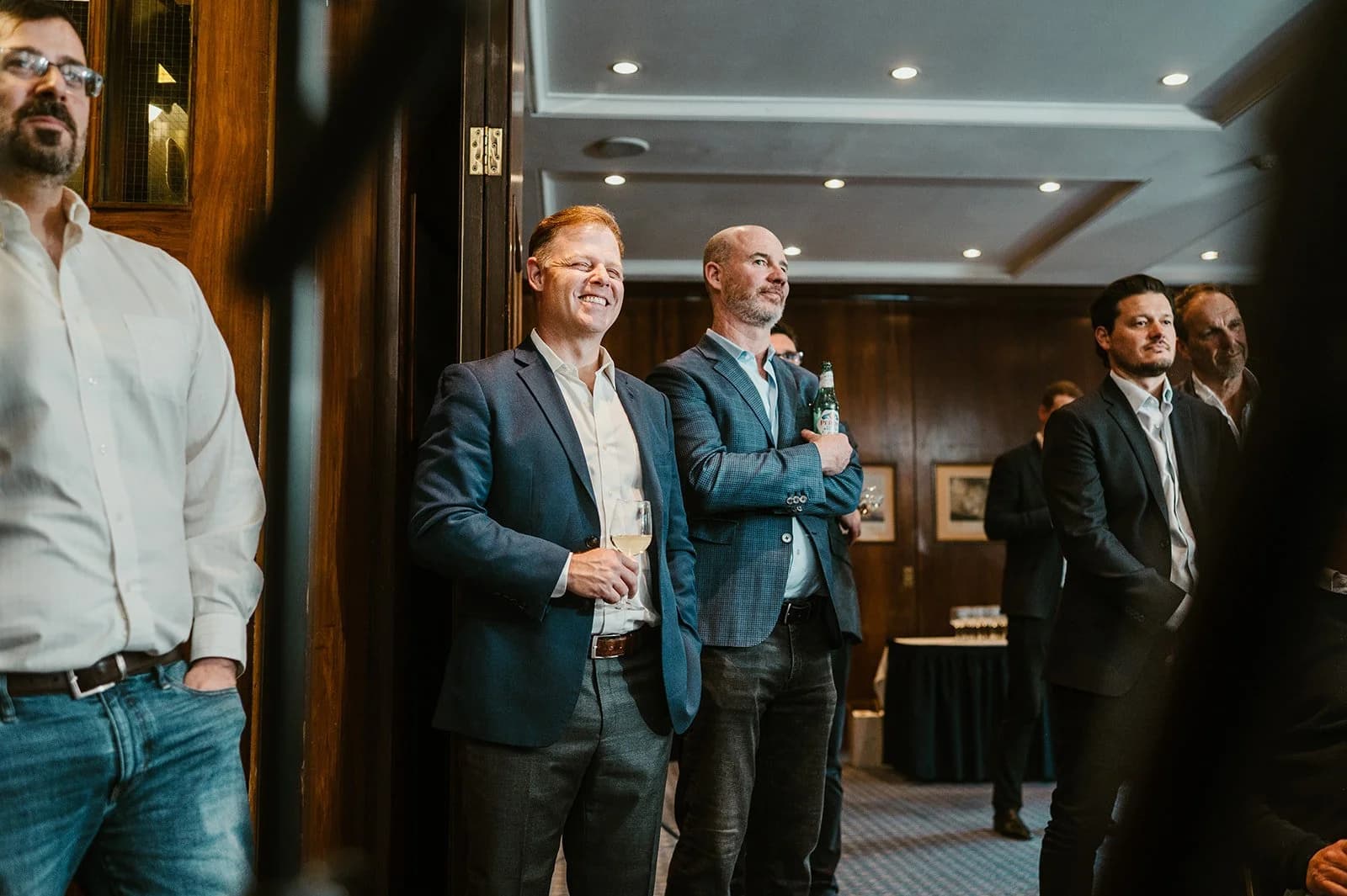 Men in blazers standing at the side of a wood-panelled room, smiling with drinks in hand