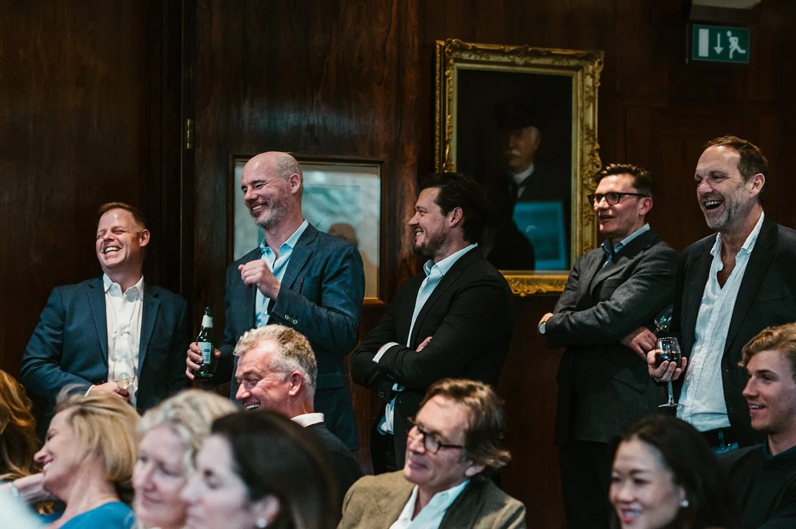 A row of guests laughing as they watch a talk, with seated listeners below them