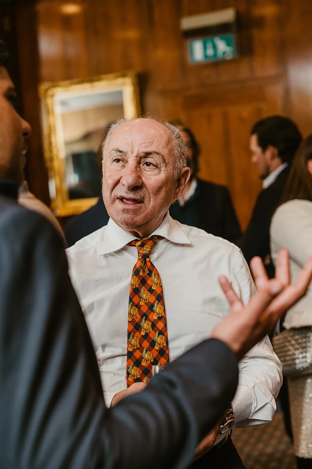 A man in an orange checked tie mid-conversation, a guest gesturing in the foreground