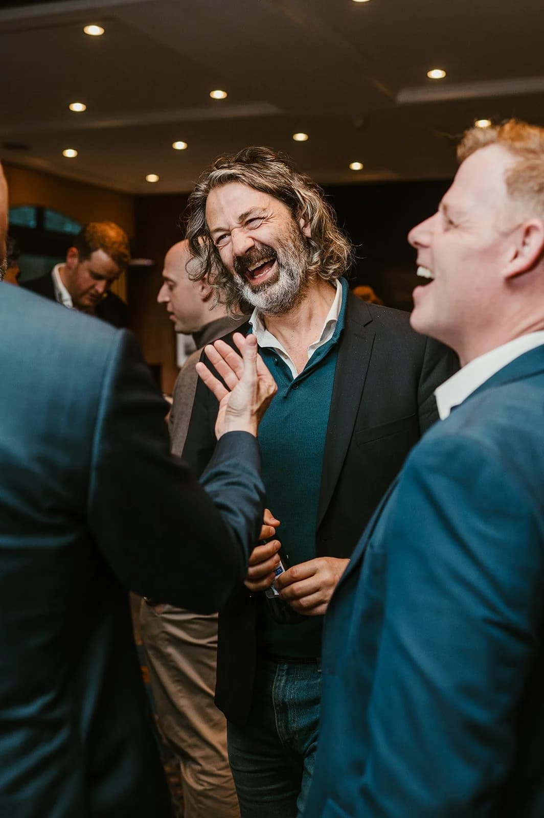 A bearded man with wavy hair laughing among other guests at a busy reception