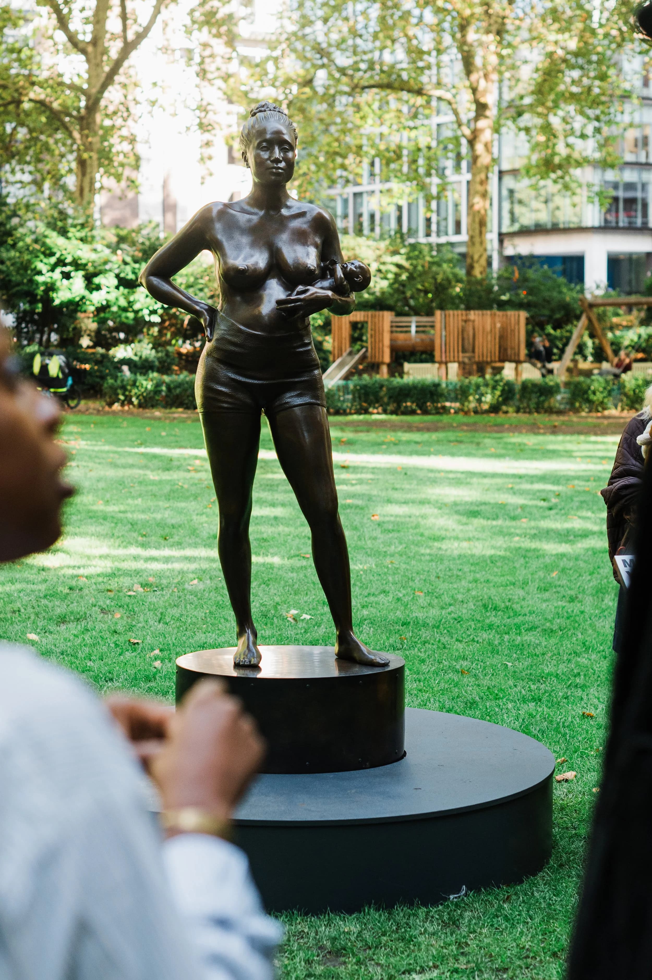 A bronze statue of a standing woman holding a baby on a plinth in a leafy London square