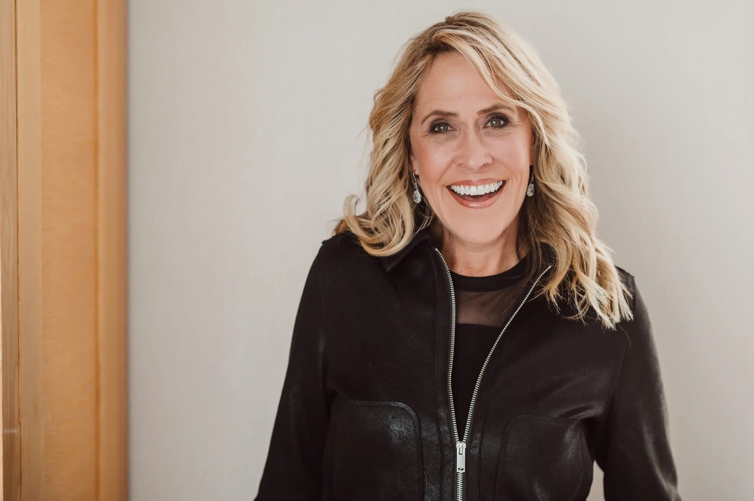 A smiling woman with wavy blonde hair in a black leather jacket beside a wooden door