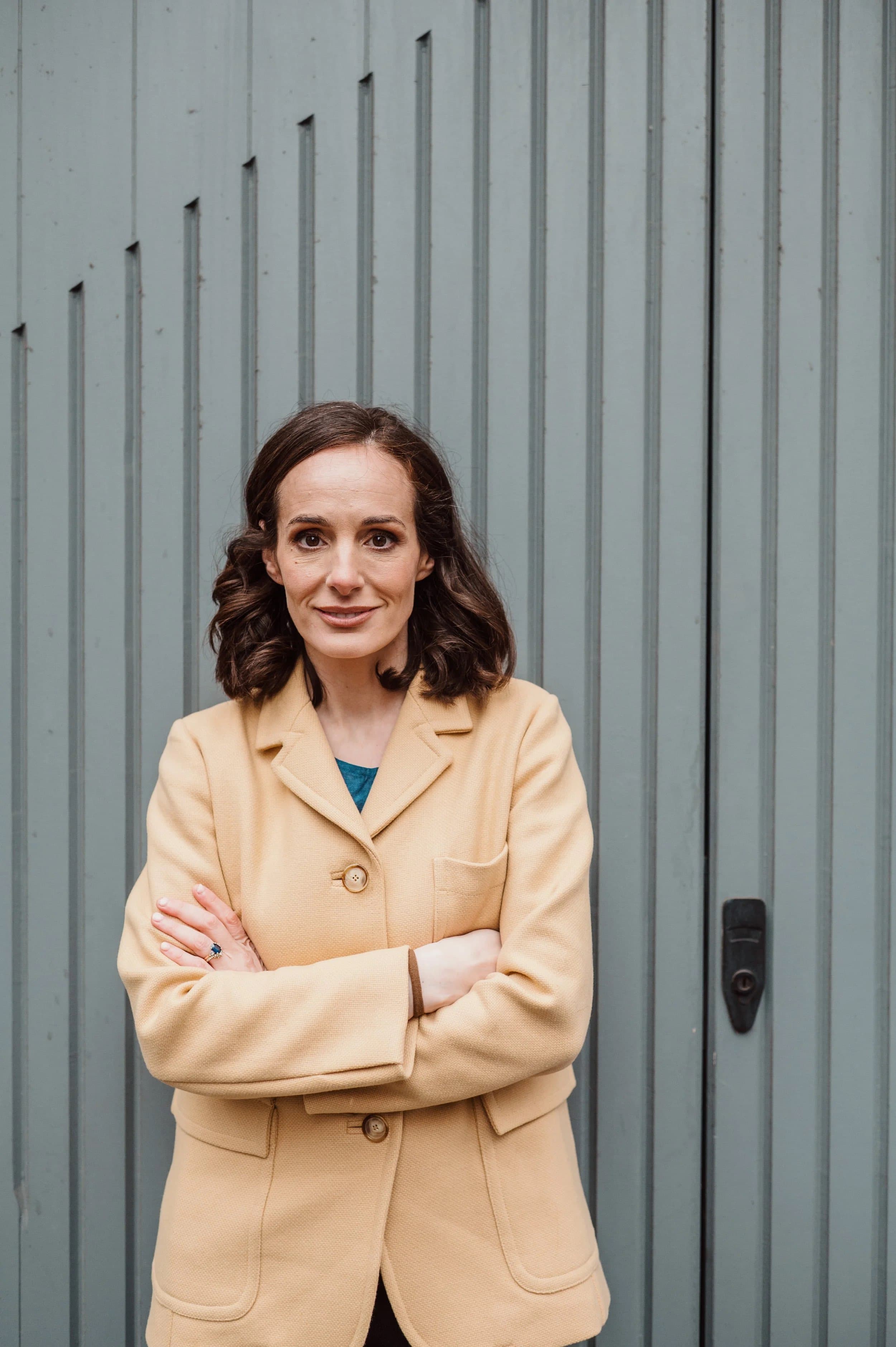 A woman in a camel jacket with arms folded in front of a grey panelled door