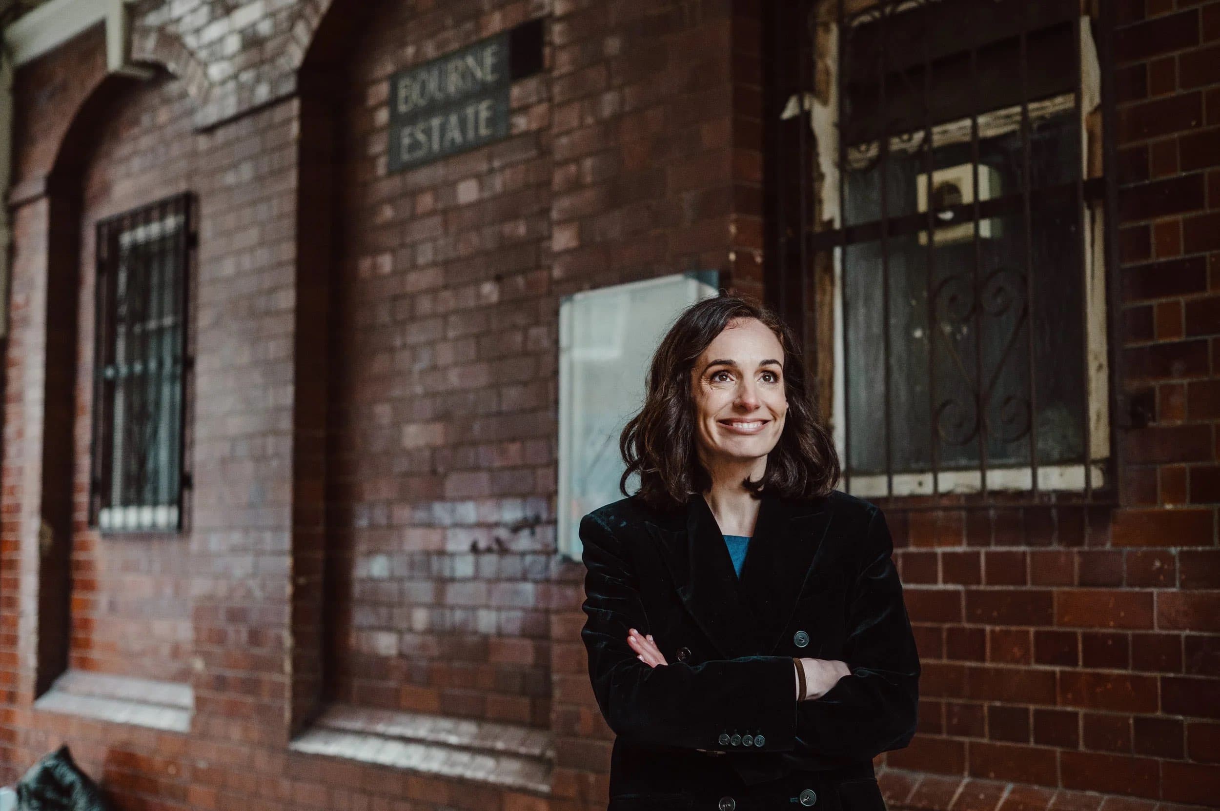 A woman in a black coat with arms folded outside a red-brick building